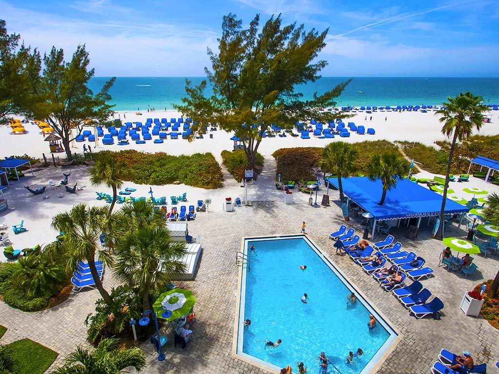 Beachfront resort scene with pool, beach, and ocean. Blue chairs, sky, and water. People swimming and sunbathing.