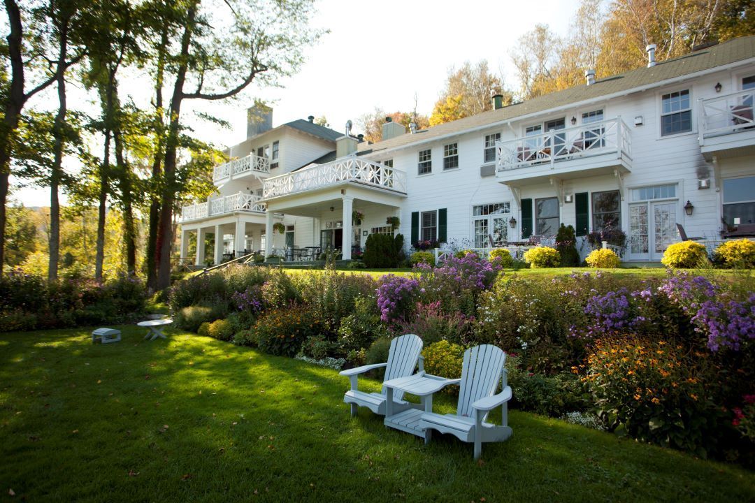 White house with porch and balcony, surrounded by a sunlit green lawn, flowers, and two white chairs.
