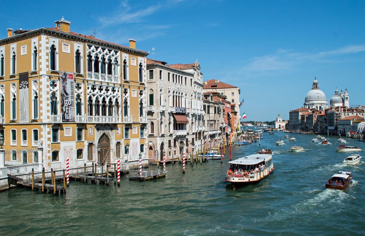 Venice Grand Canal with boats, buildings, and the Santa Maria della Salute church under a bright blue sky.