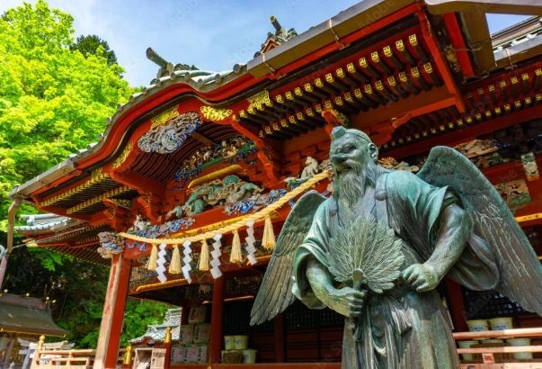 A bronze statue of a winged Tengu holding a fan in front of a red, ornate Japanese temple building with green trees.