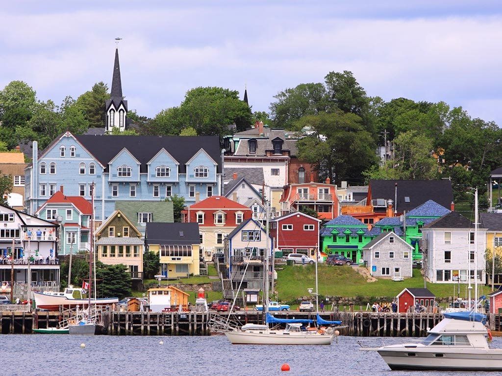 A colorful waterfront town with houses, a church steeple, and boats docked at a pier under a cloudy sky.