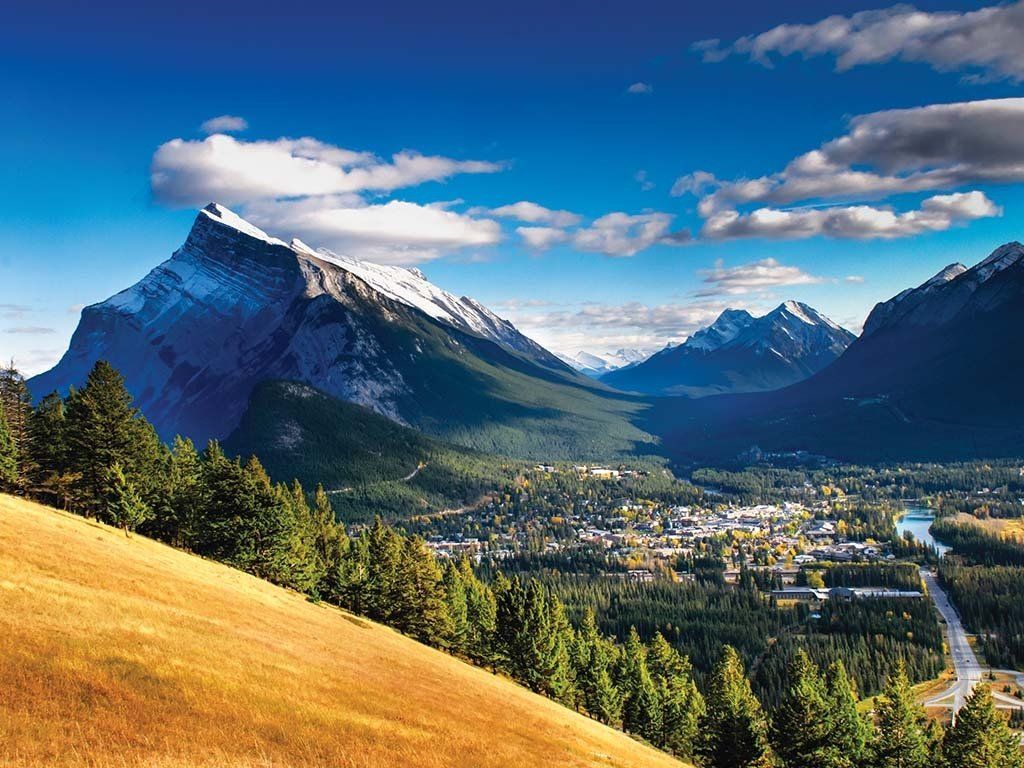 Sunny mountain valley with a snowcapped peak, forest, town, and blue sky with clouds
