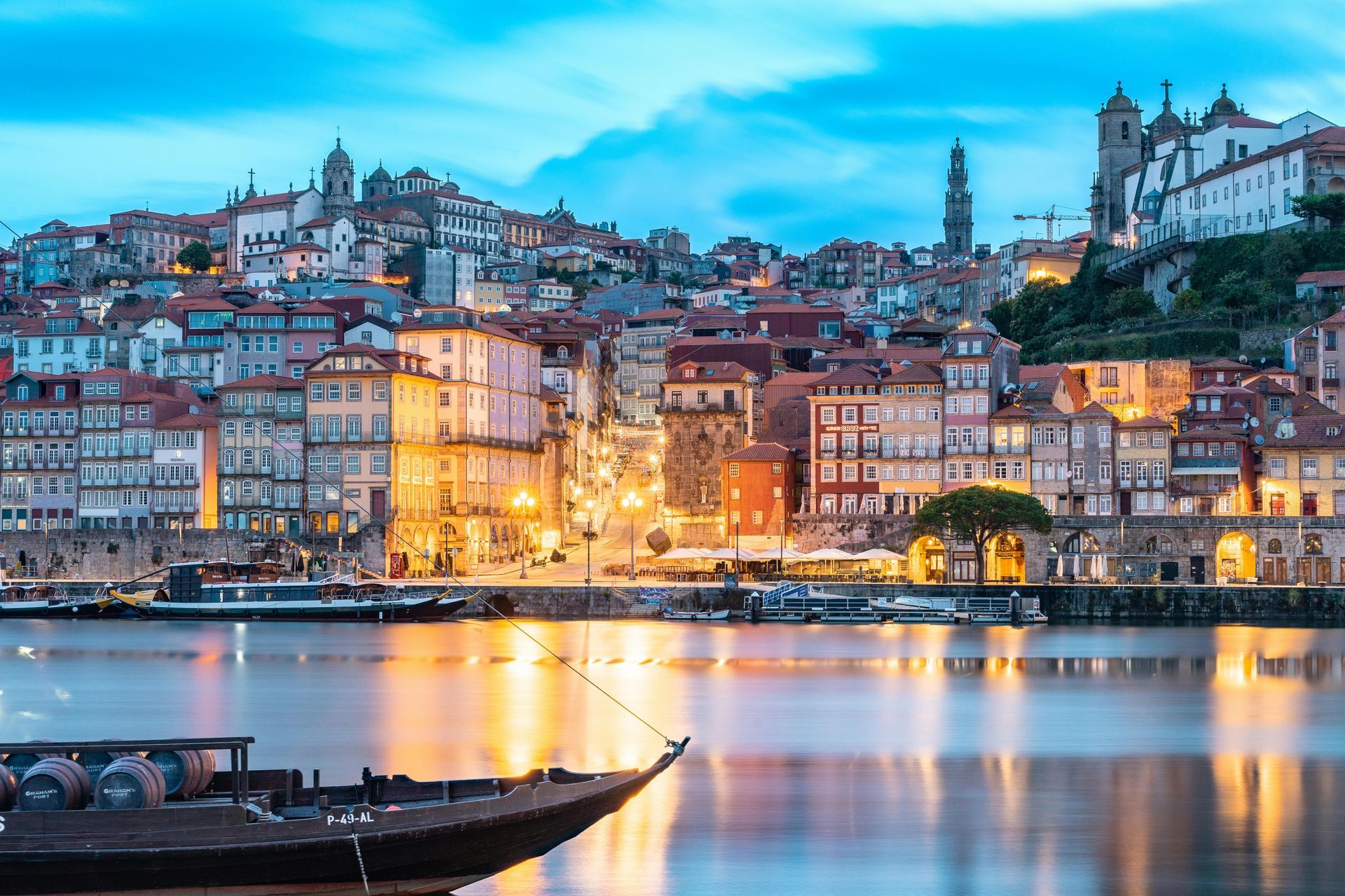 Dusk harbor view of a colorful hillside city with lit buildings reflected on calm water, boat in foreground
