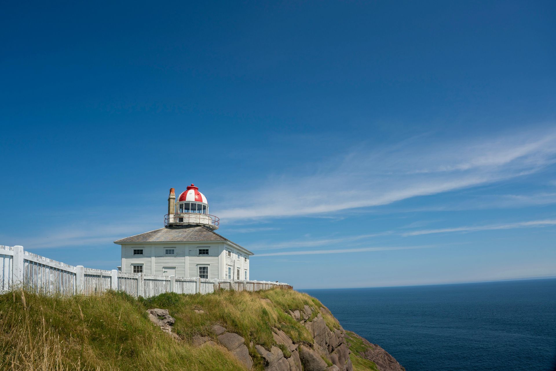 White lighthouse with a red-and-white dome on a cliff, overlooking the blue ocean under a clear blue sky.