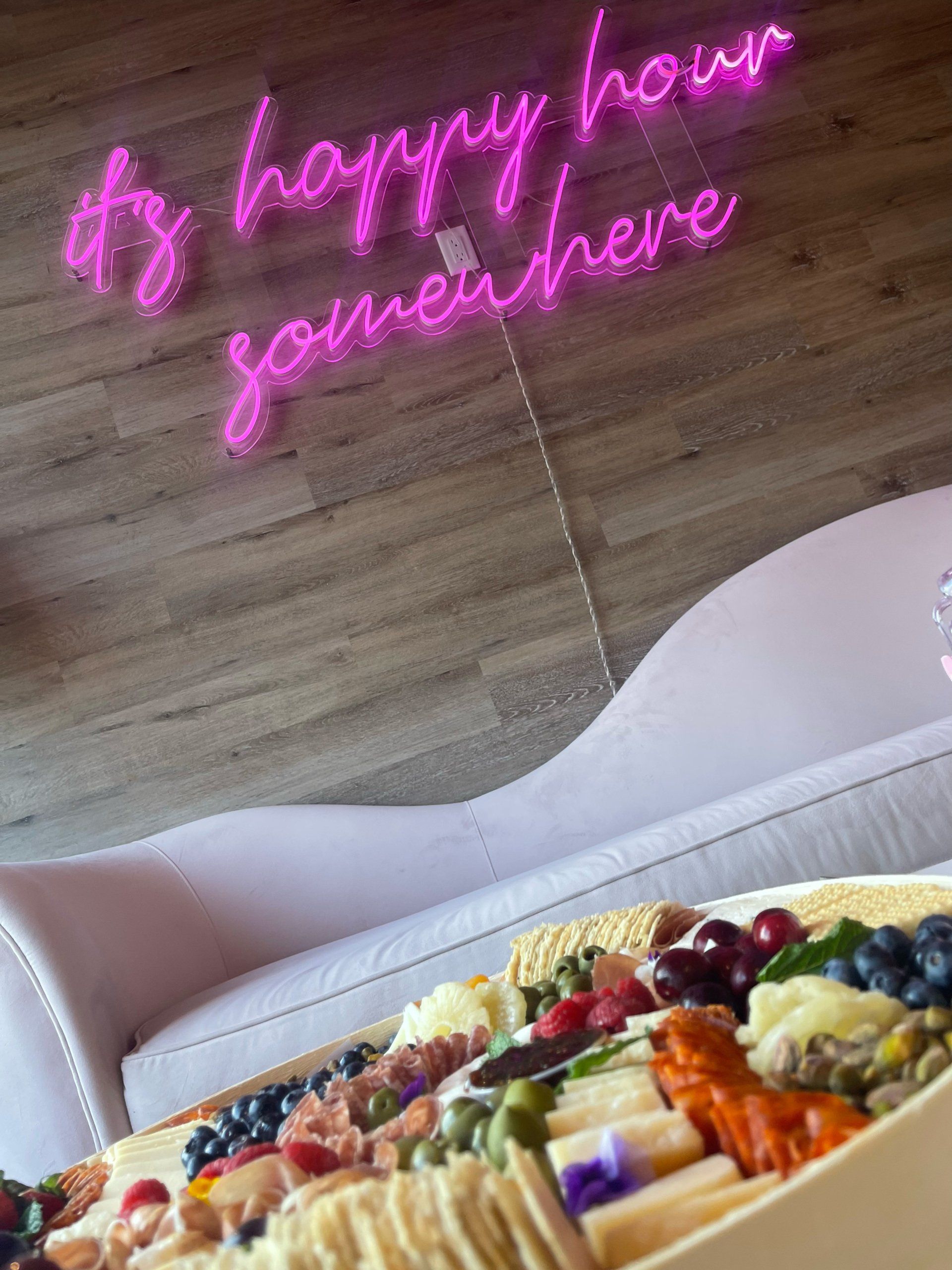 A pink neon sign that says `` it 's happy hour somewhere '' is above a tray of food.