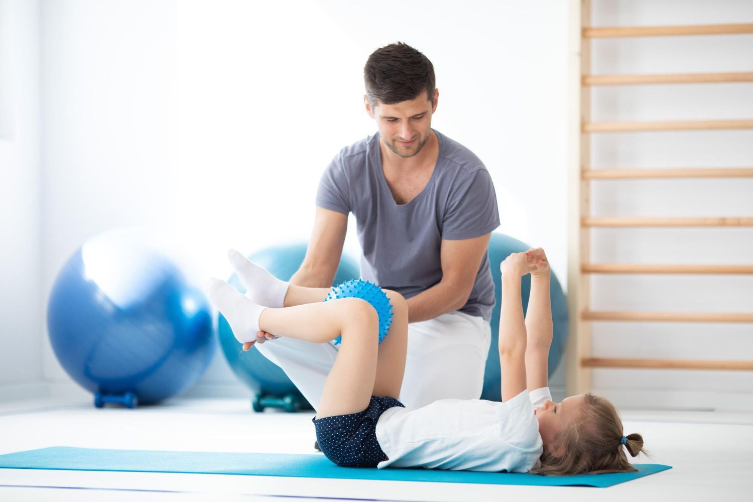 Man assisting a young girl doing physical therapy on a mat. Blue balls and wall bars in the background.