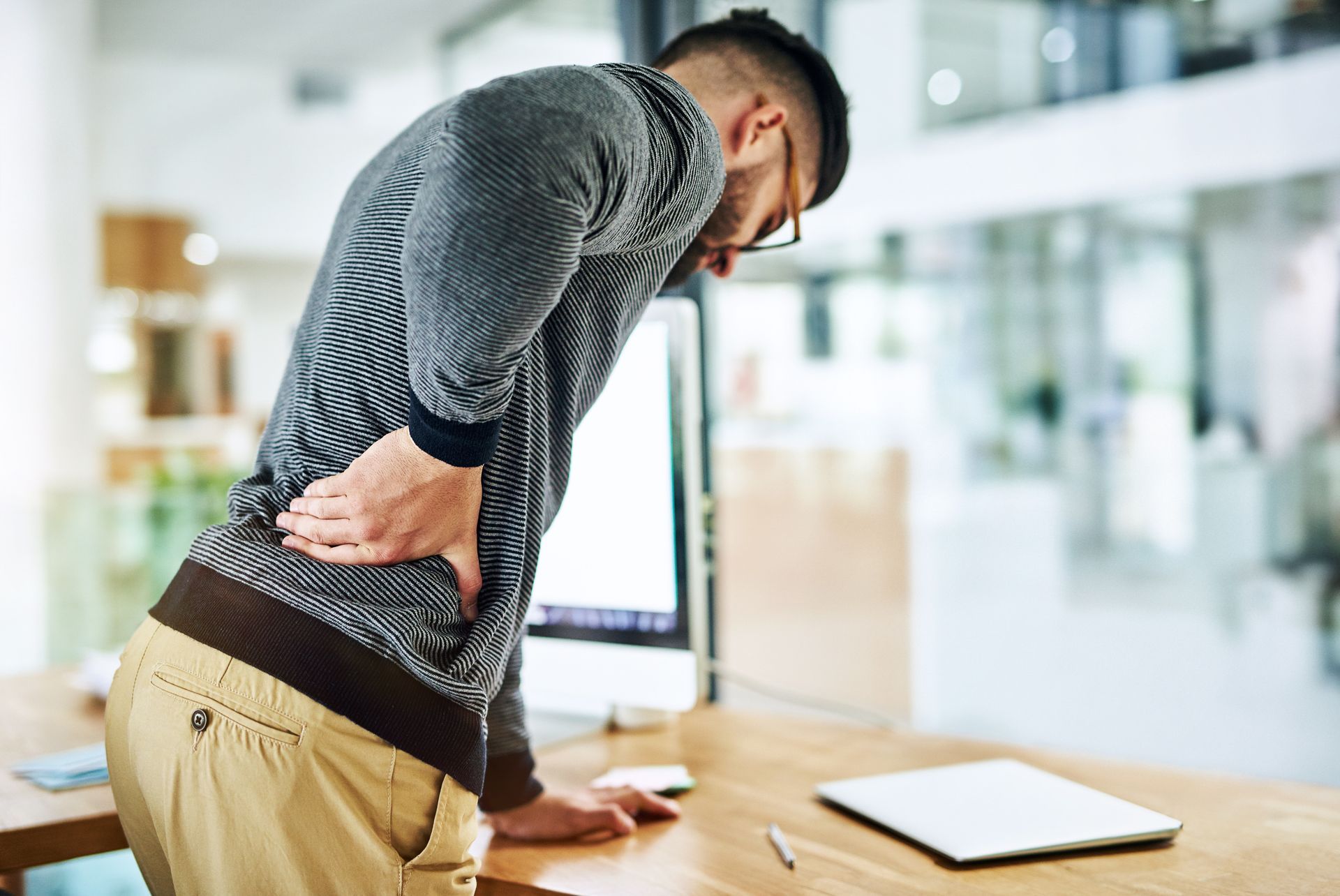A man is standing at a desk holding his back in pain.