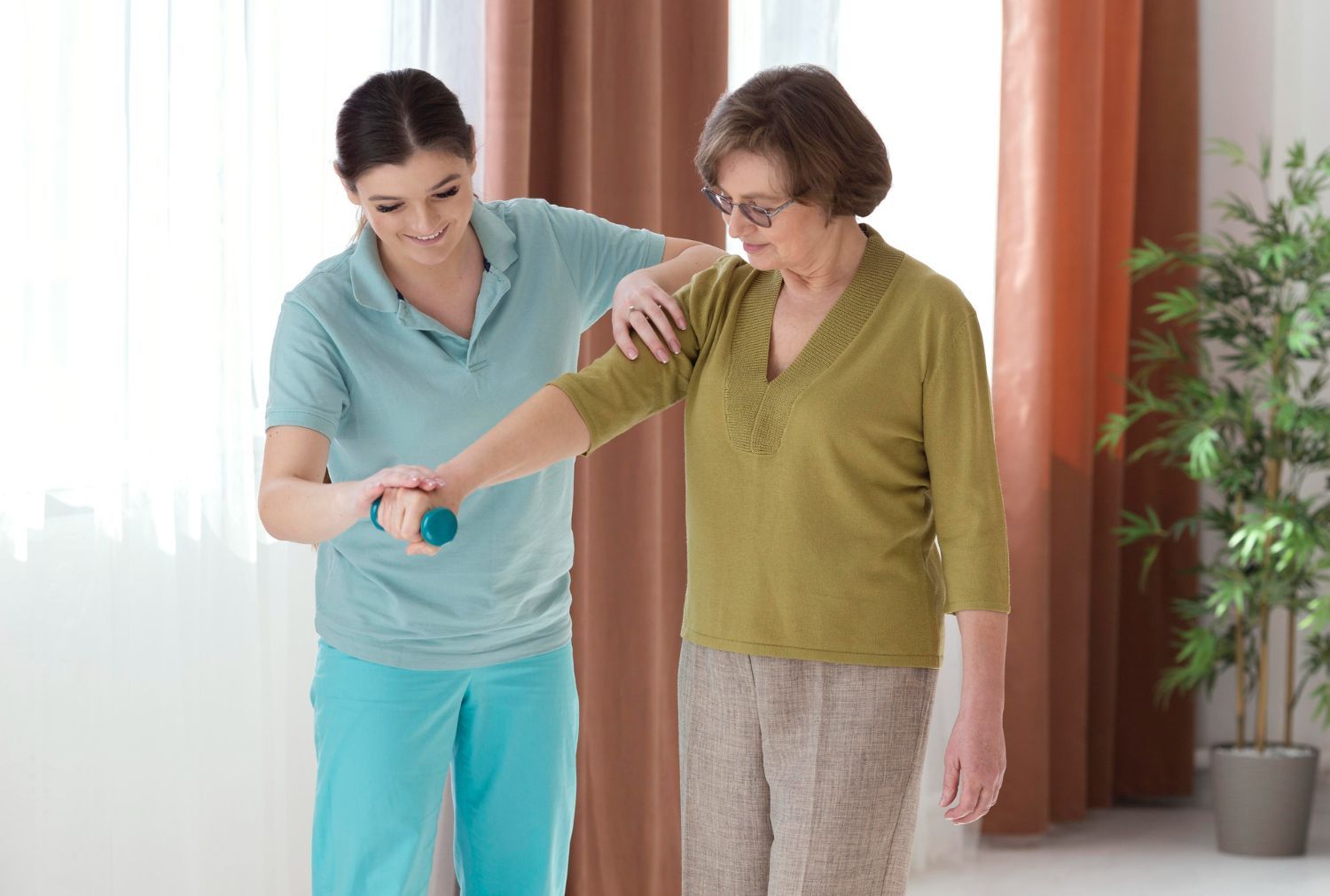 Woman helps an elderly woman with arm exercise, indoor setting.