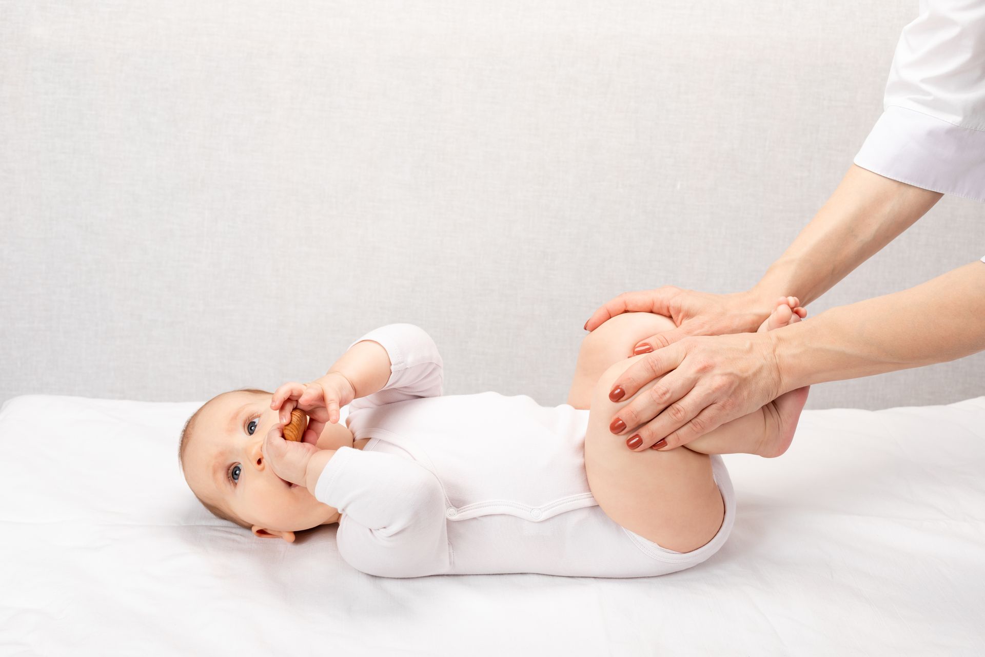 Baby in white onesie has feet held by unseen hands. Baby holds object to mouth.