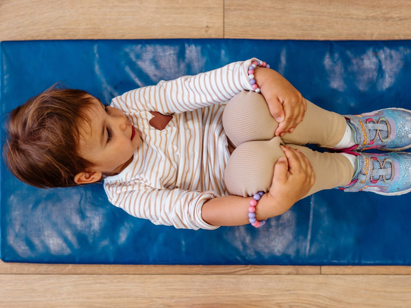Child on blue mat, knees to chest, looking left. Light-colored outfit, tan socks, and patterned shoes.