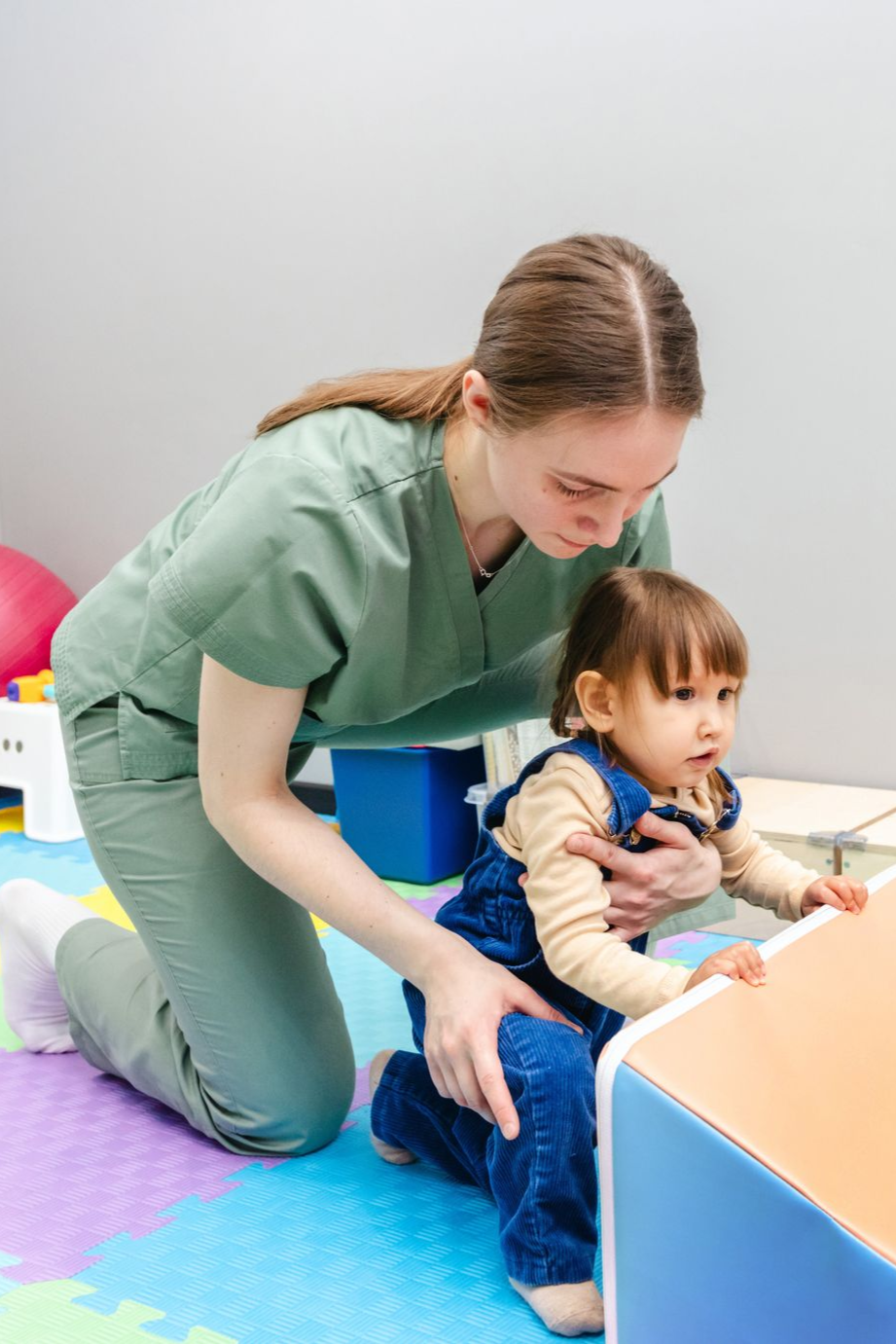 Therapist assists toddler with walking, hands on chest and leg, in a therapy room.