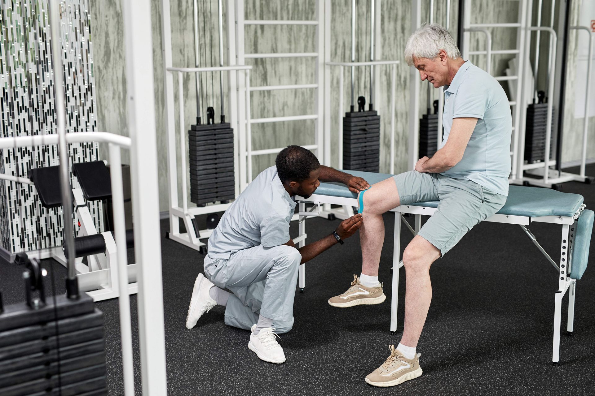 Therapist examining patient's knee in a rehab room. Both are wearing light-colored clothes.