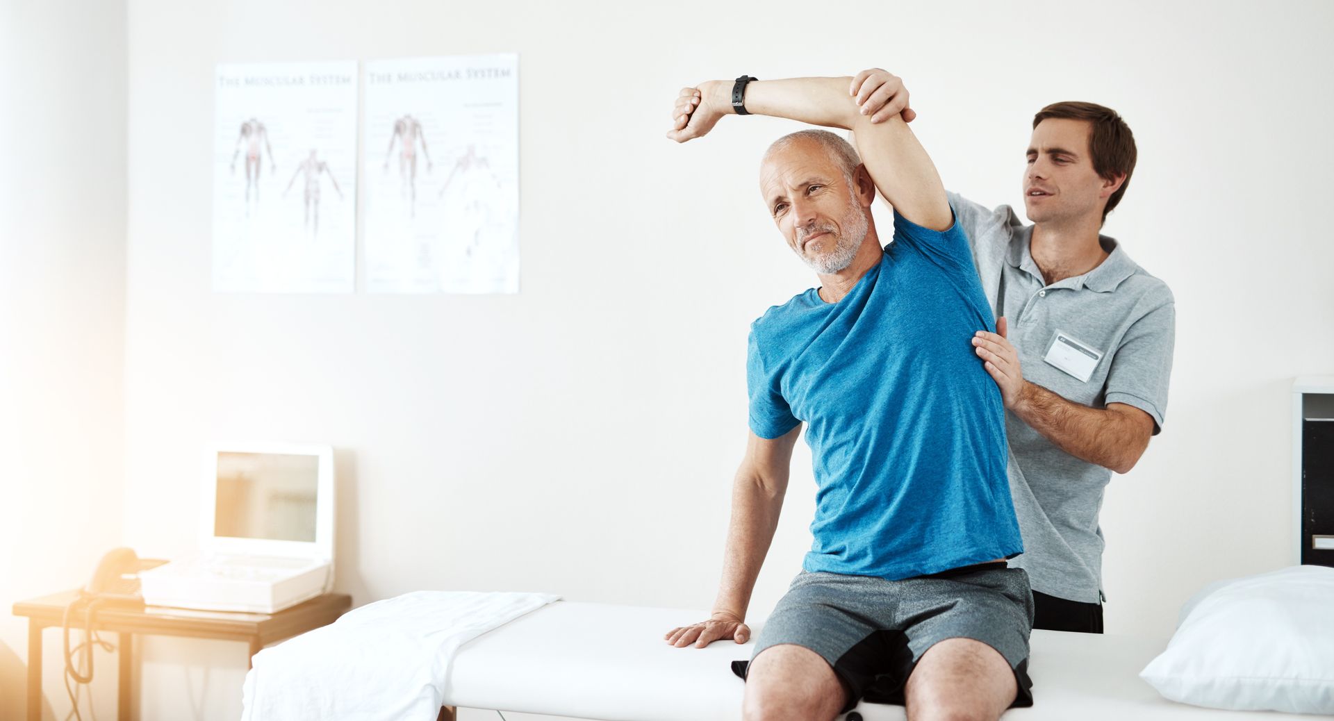 Therapist assisting a patient's arm stretch in a white room. The patient is wearing blue, and the therapist is wearing gray.