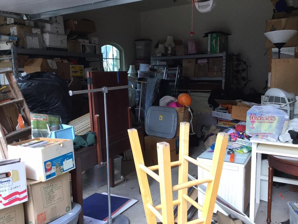 Cluttered garage interior with boxes, furniture, and various items, including a step stool and a garment rack.