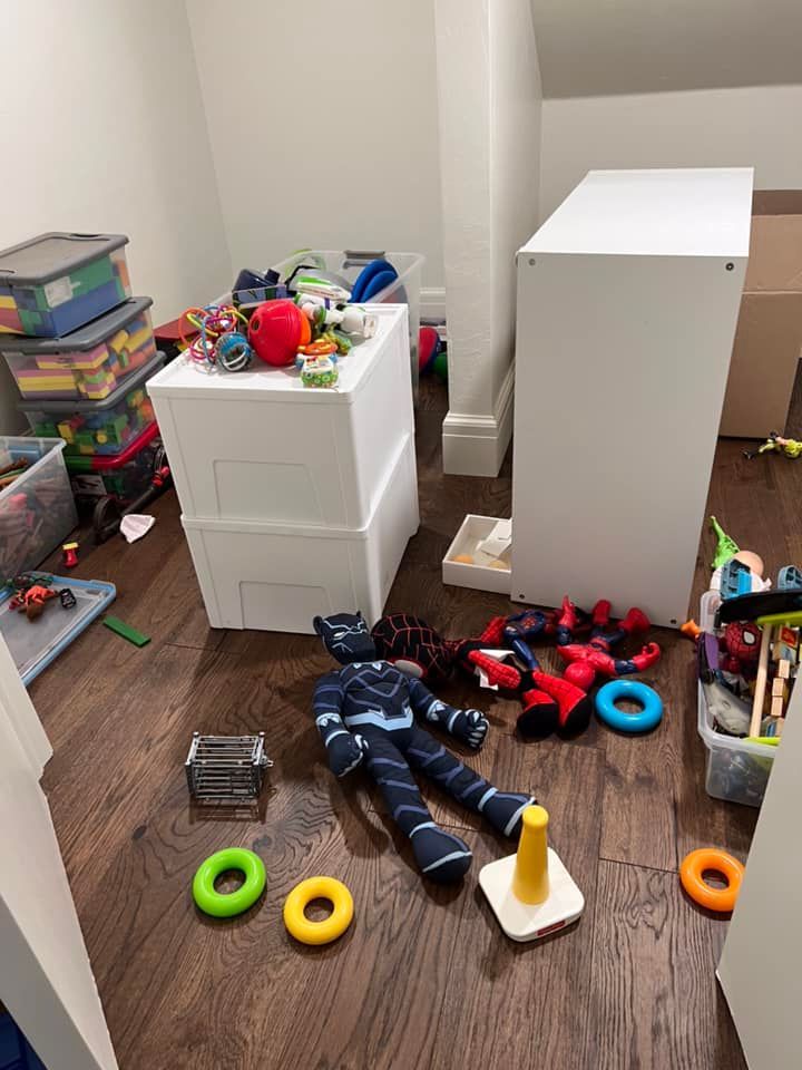 Messy playroom with toys scattered on the wooden floor. Two white storage units stand near a wall.