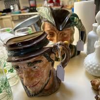 Two ceramic beer mugs. One is an old man's face, the other is a sailor's head. They sit on a white table.