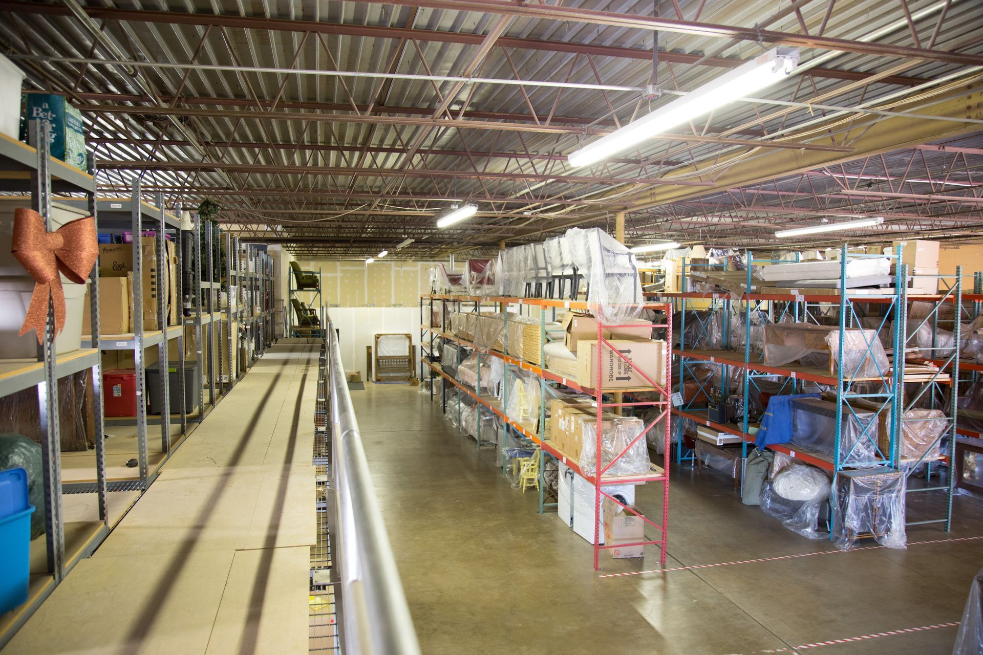 Warehouse interior with metal shelving holding various boxes and items. Fluorescent lighting illuminates the space.