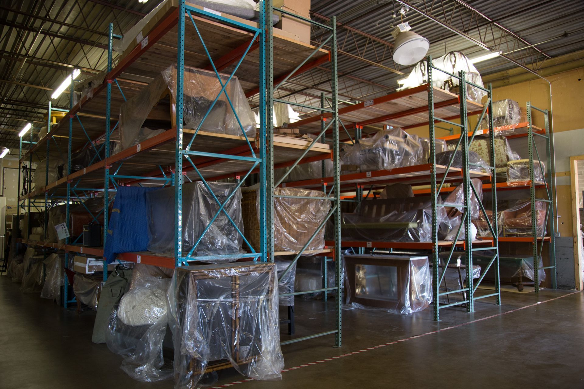 Warehouse shelves filled with plastic-wrapped furniture and equipment. Industrial setting with fluorescent lighting.