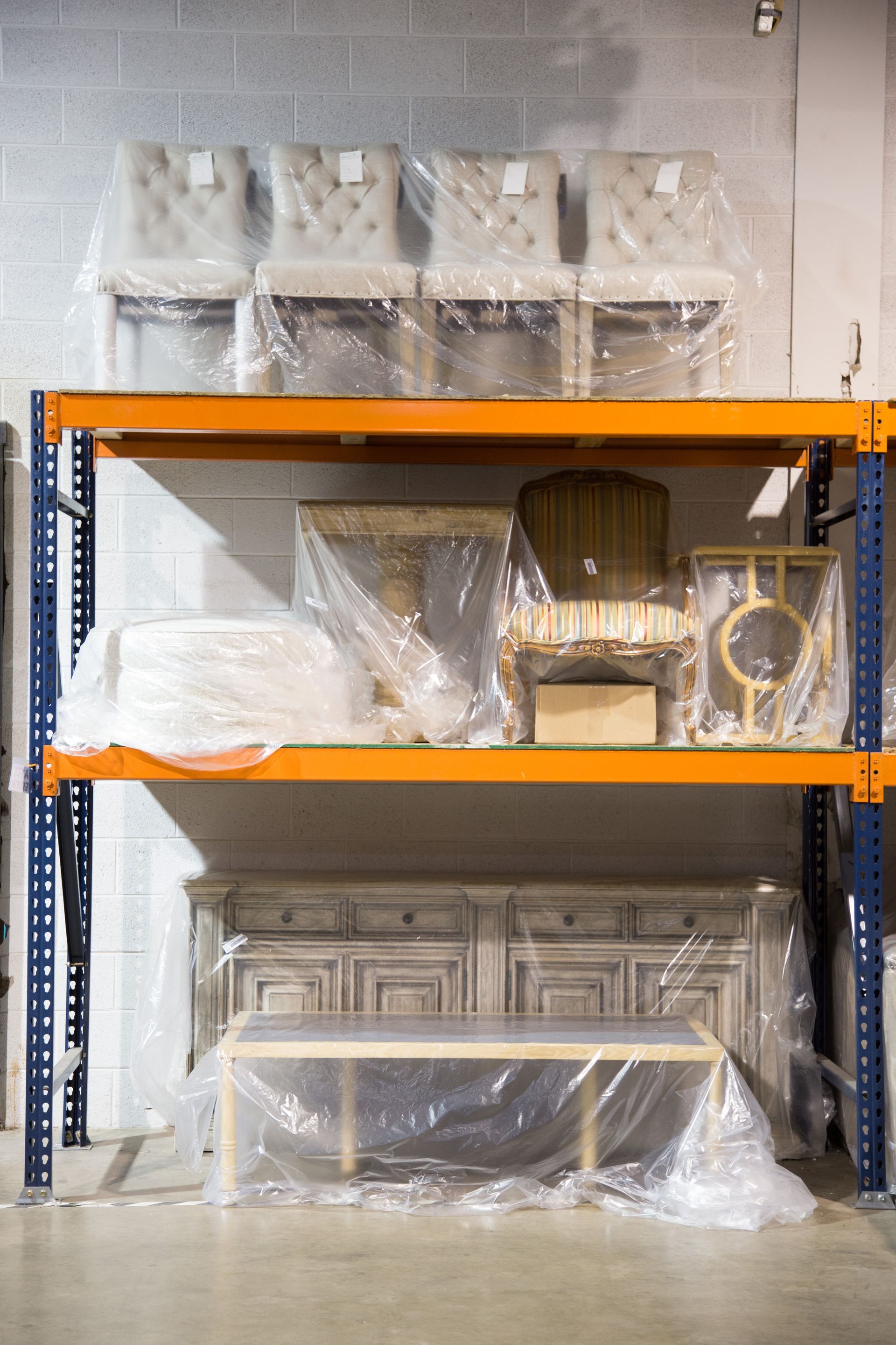 Furniture on shelving in a warehouse, wrapped in plastic. Chairs, a buffet, and a table are visible.