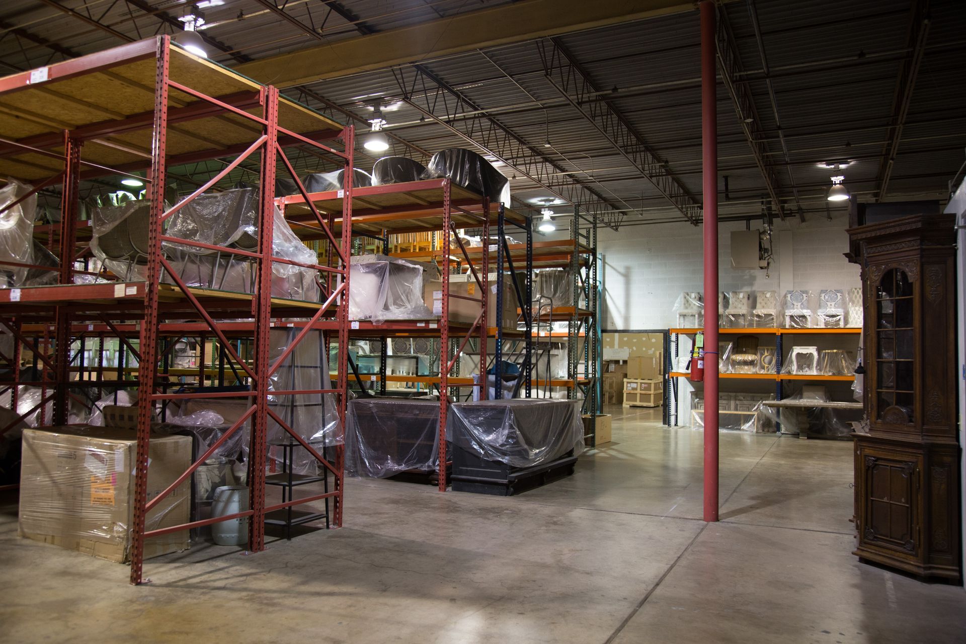 Warehouse interior with metal shelving filled with packaged items. Large brown cabinet to the right.