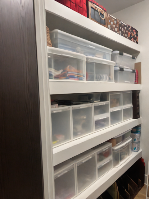 White shelves filled with clear plastic bins, boxes, and a few decorative containers in a closet.