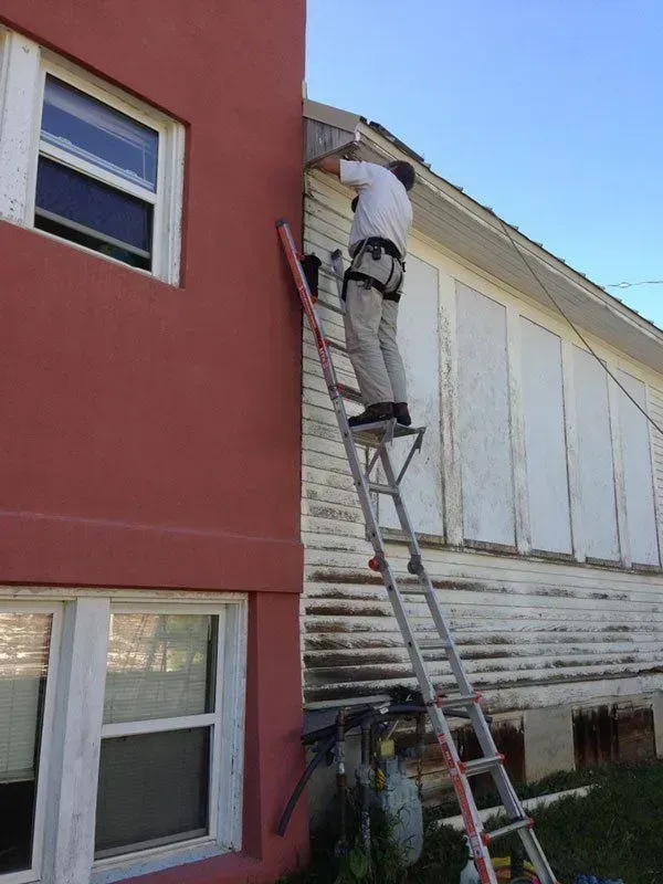 A man stands precariously on an extended ladder, working on the exterior wall of a house. 