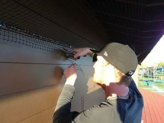 A person in a cap works diligently, installing a net on a wooden structure. 