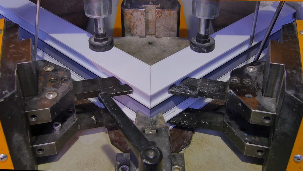 Macchina metallica che unisce due pezzi di plastica bianca in un angolo.