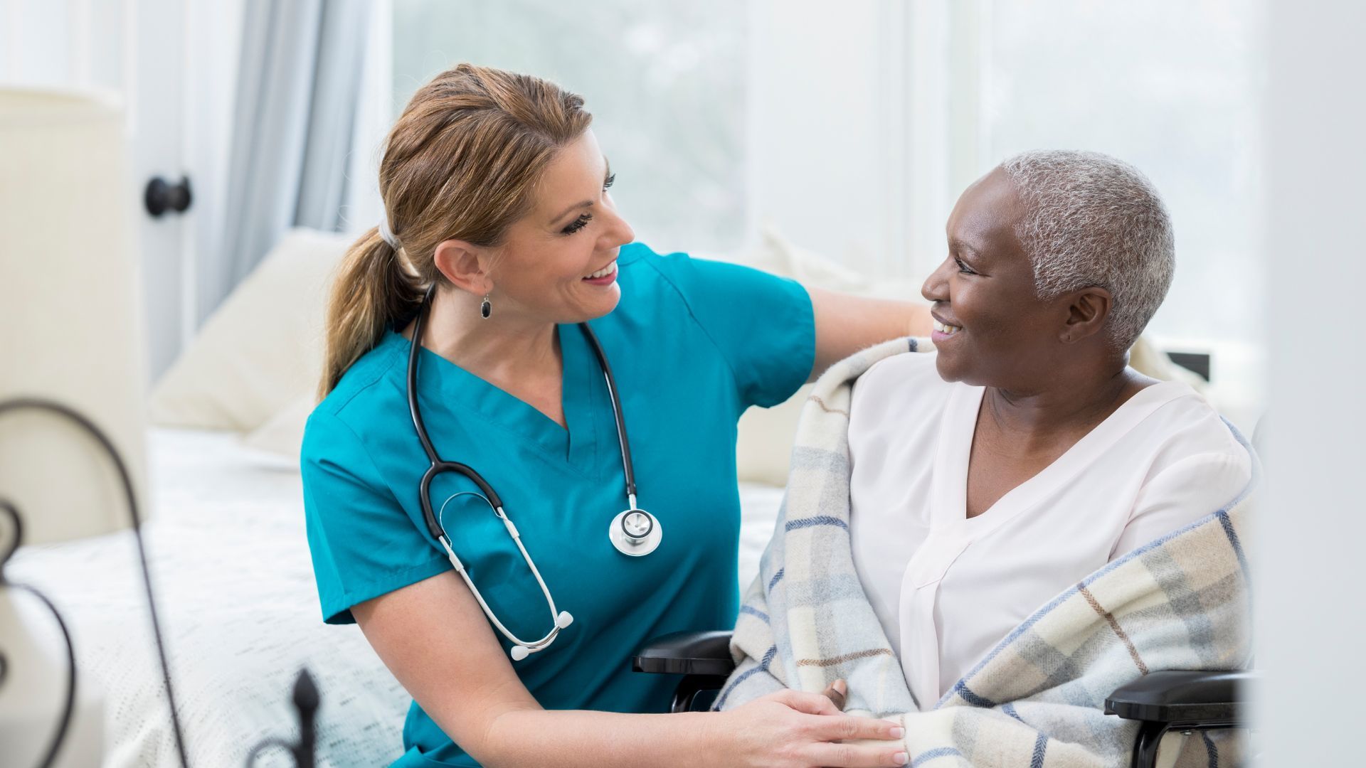 A healthcare worker in scrubs smiles at an elderly person seated in a wheelchair; indoor setting.