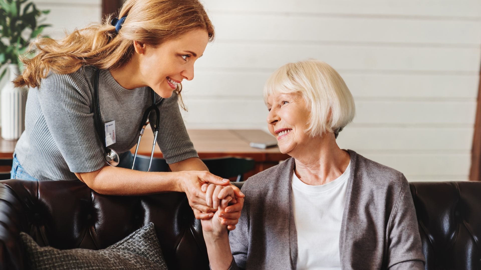 Woman holds the hand of an older person, both smiling, in a home setting.