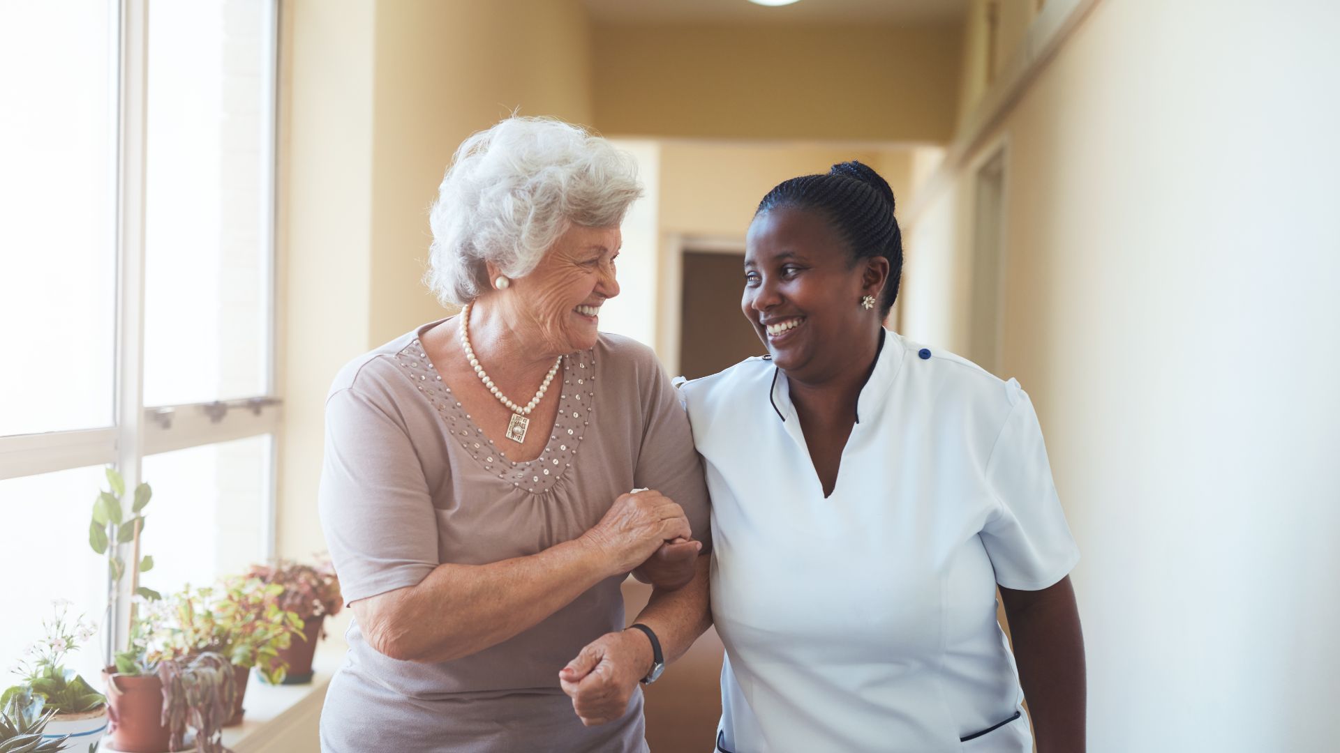 Woman helping another woman walk down hallway, both smiling.