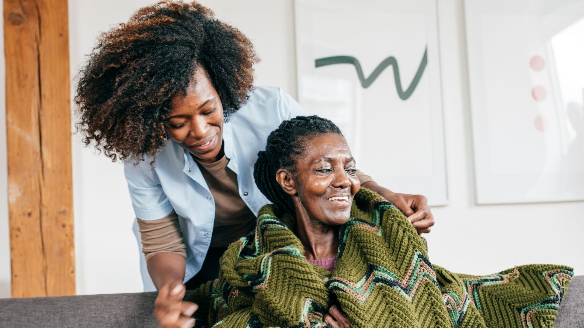 Woman covers another woman with a blanket, both smiling, indoor setting, abstract artwork.