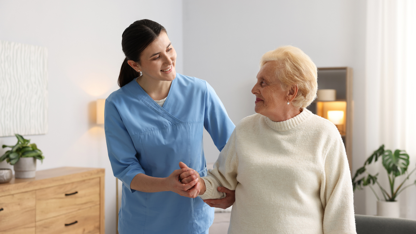 Caregiver in blue uniform assists elderly person walking indoors, smiling.