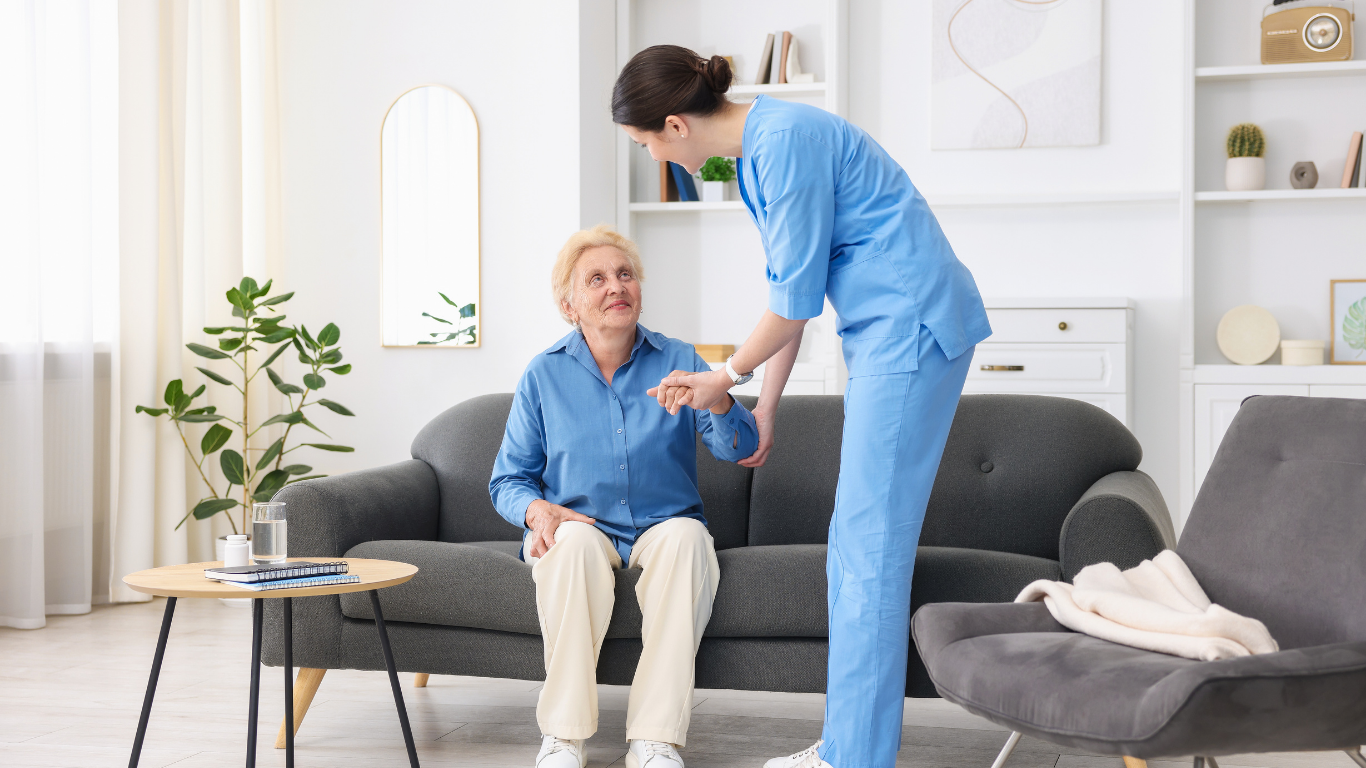Caregiver assisting an older person to stand up from a sofa in a living room.