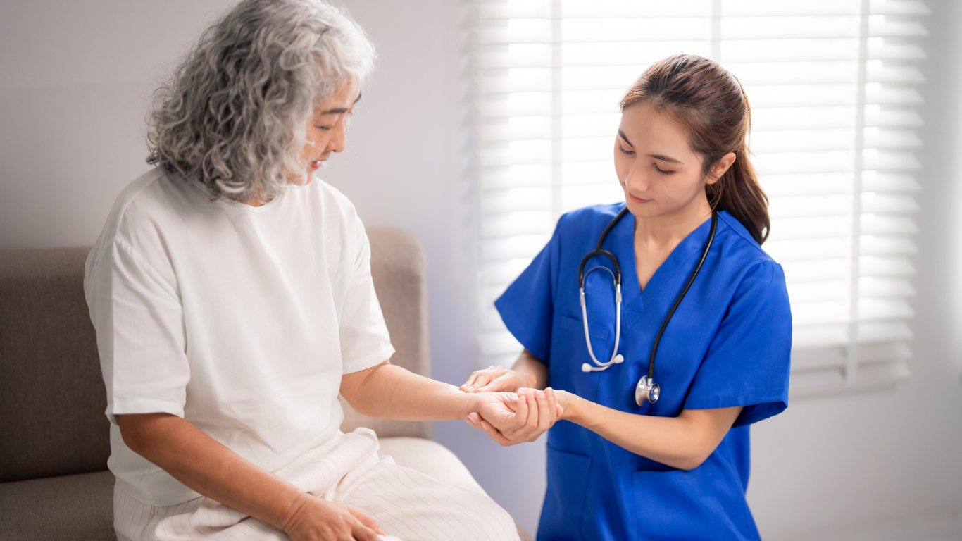 Nurse in blue scrubs examines an older adult’s wrist indoors.