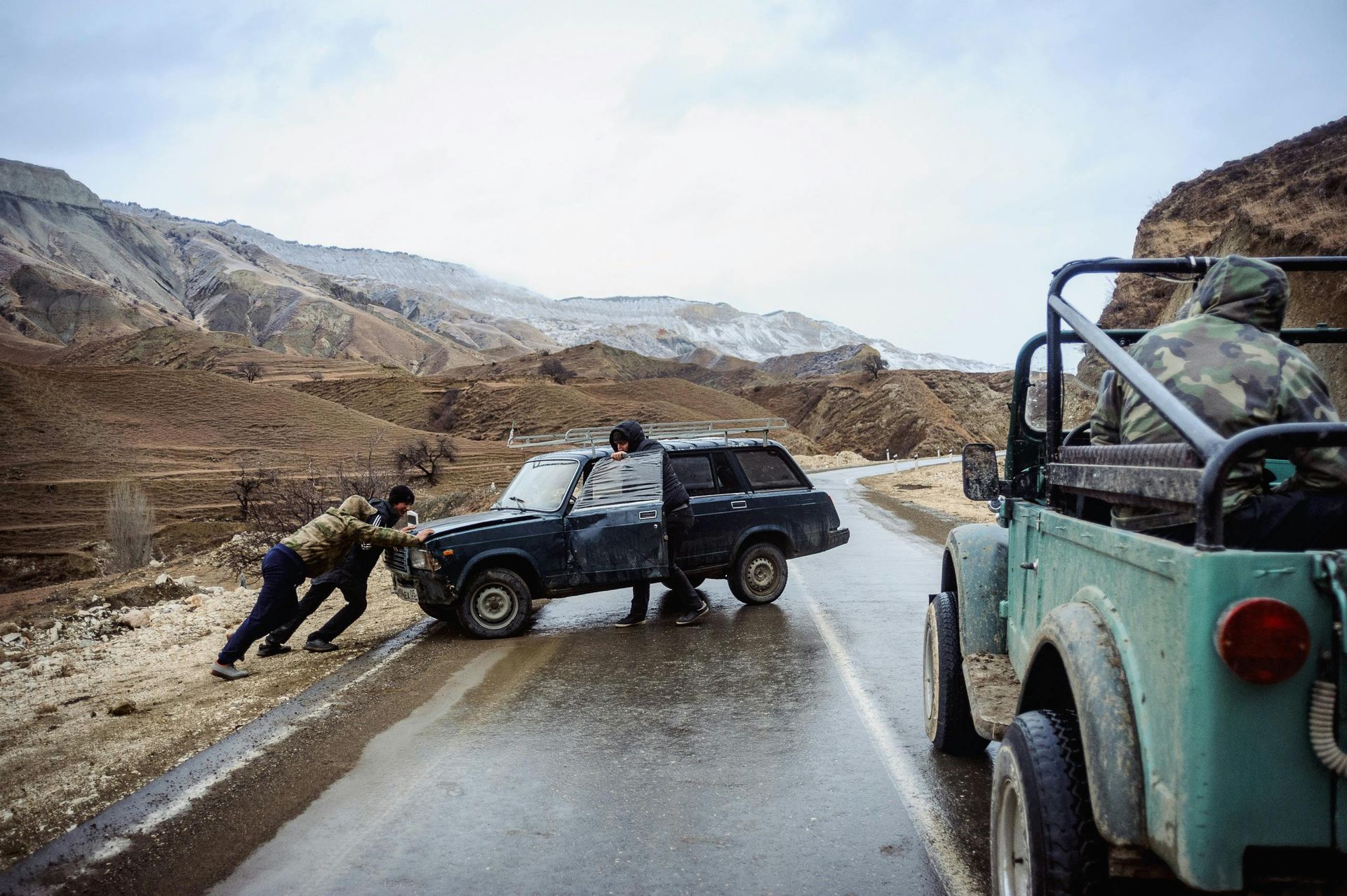 People pushing a black SUV on a wet mountain road, with a green vehicle and snowy hills nearby.