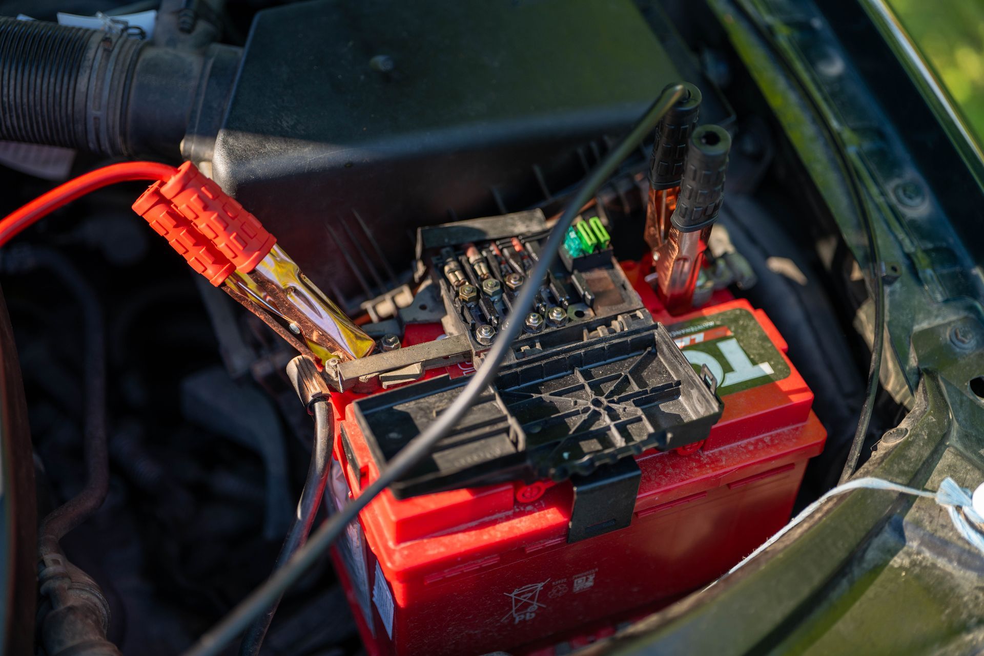 Red car battery with a battery maintainer and cables clipped on in an engine bay
