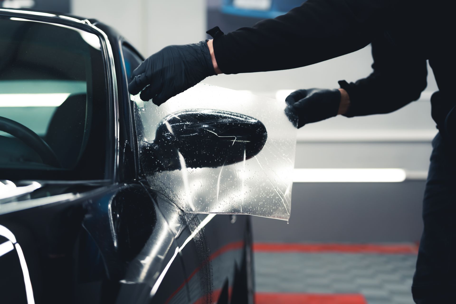 Gloved worker hand-washes a black car, spraying water on the side mirror in a garage