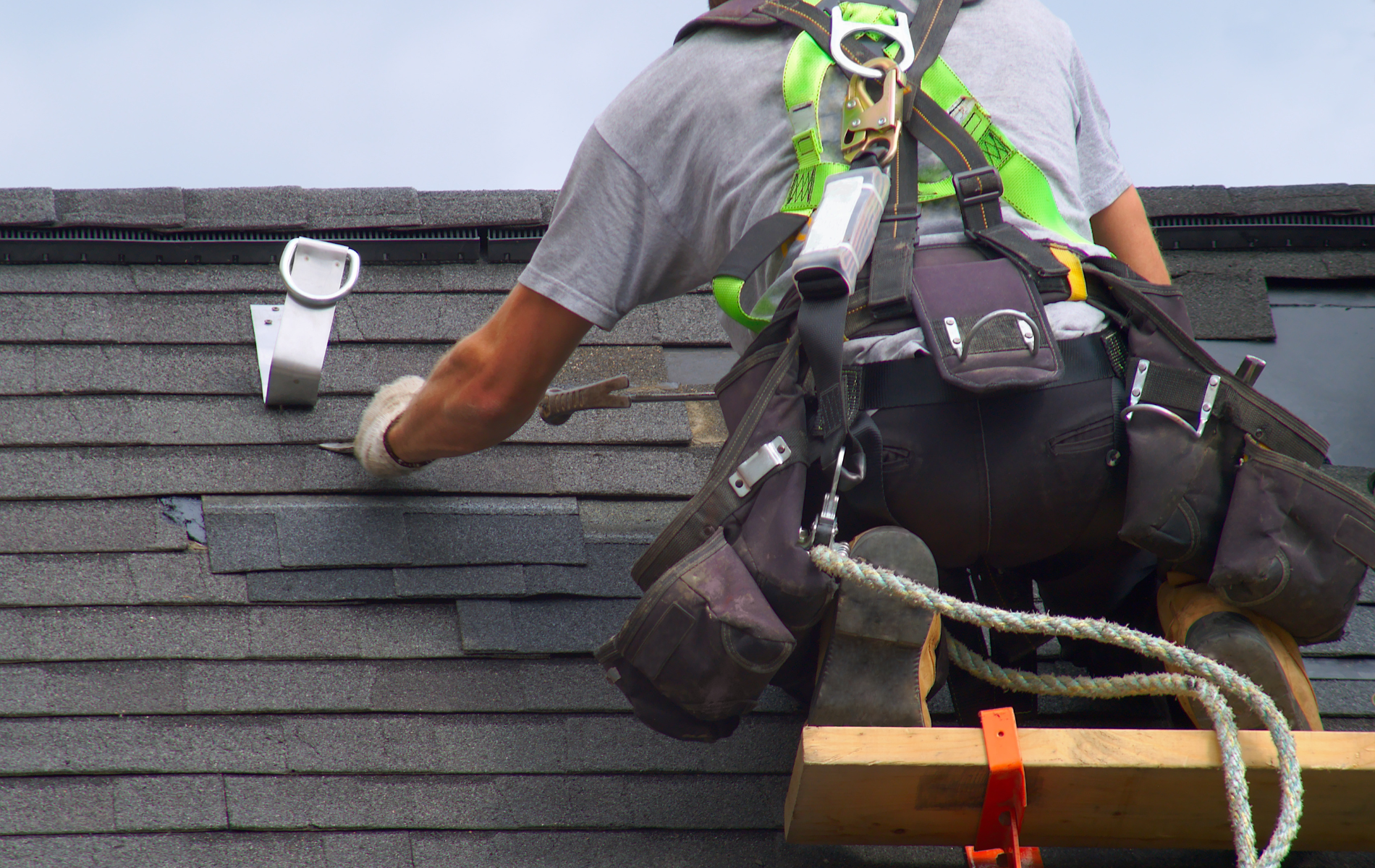 Roofer replacing shingles on a dark gray shingled roof, secured with a safety harness and tool belt.
