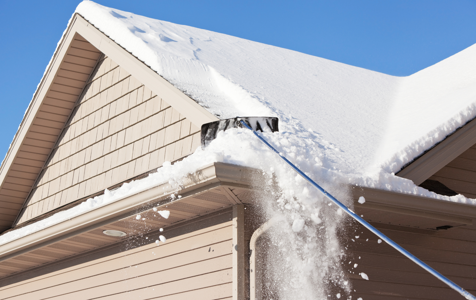 A person uses a roof rake to remove snow from a house roof on a sunny day.