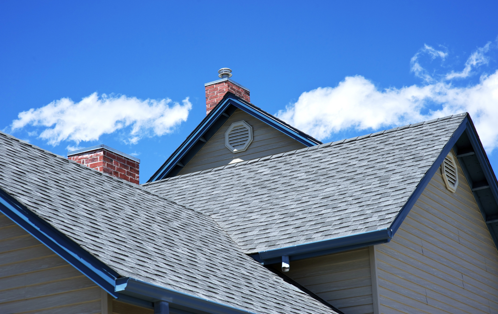 Gray shingled rooftops, two brick chimneys, beige siding, blue sky with white clouds.