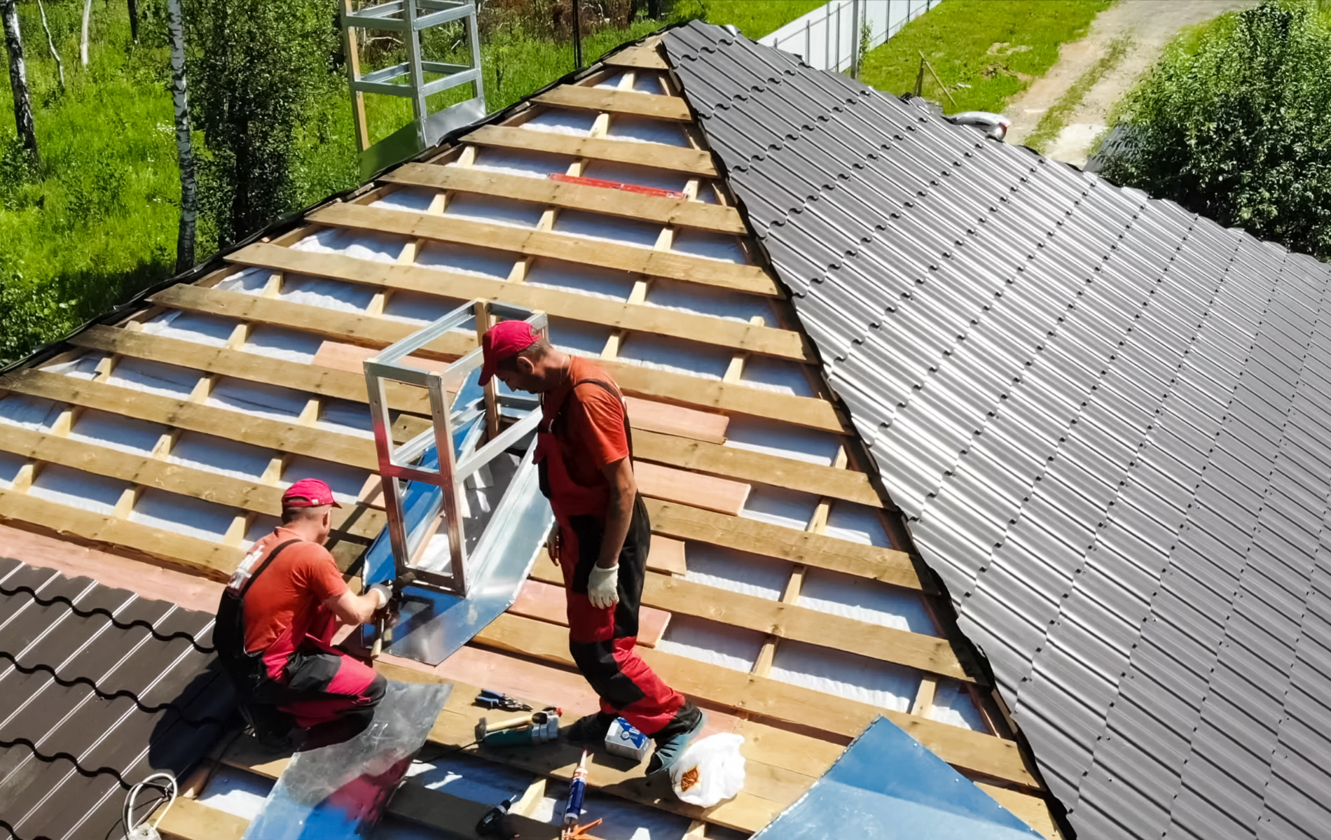 Two roofers in red overalls installing metal roofing on a house.
