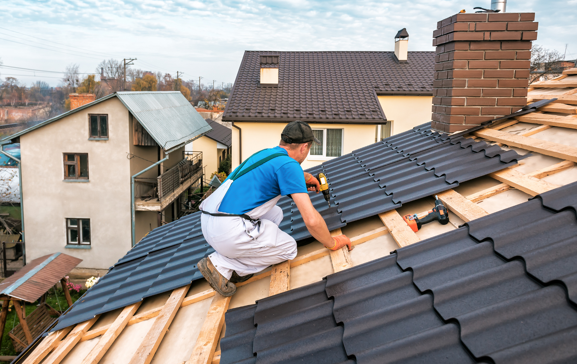 Roofer kneeling, installing dark metal roof tiles on a house. Other buildings and chimney visible in the background.