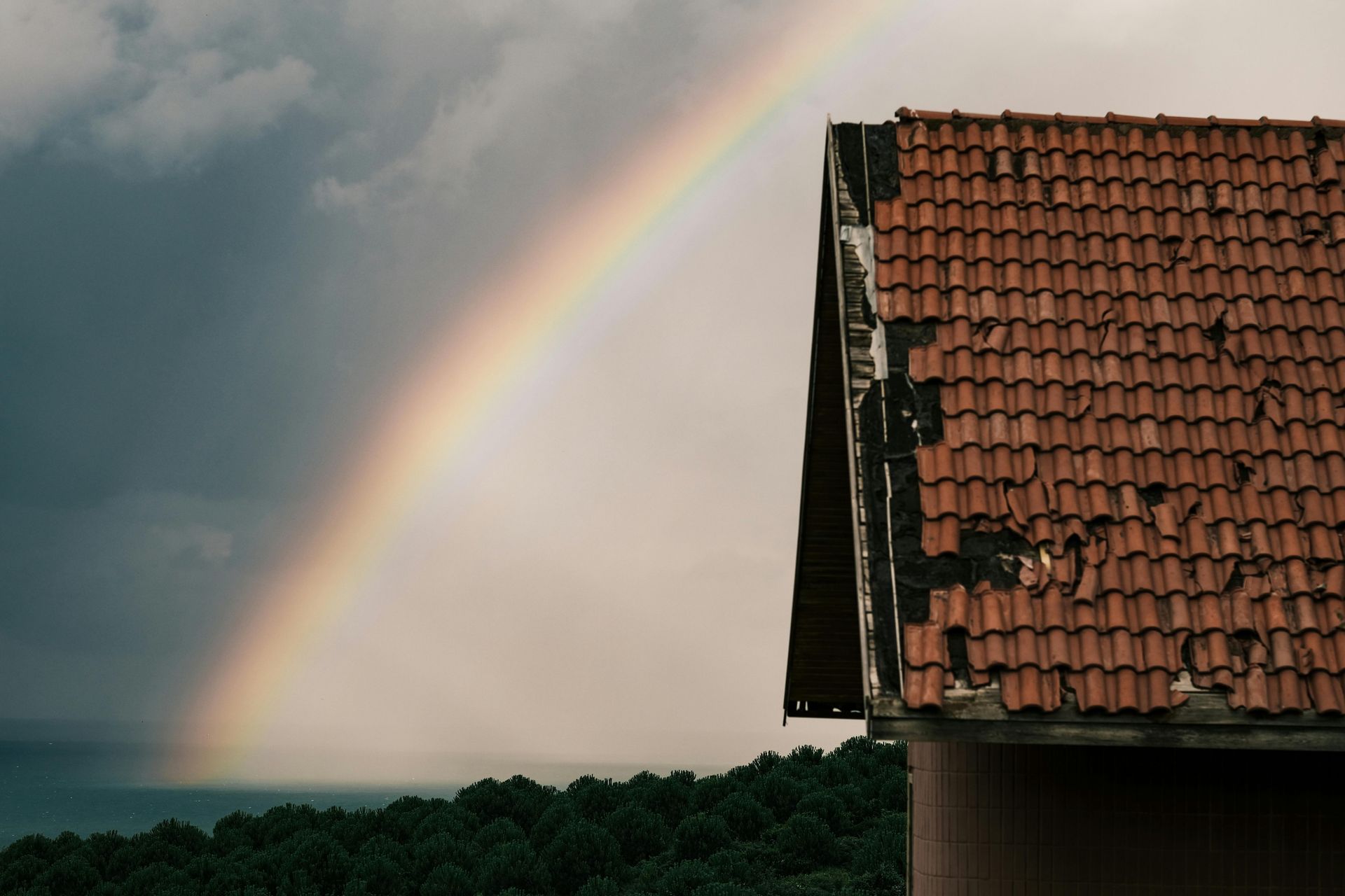Rainbow arches over a house with a weathered red-tiled roof, near a dark forest and cloudy sky.