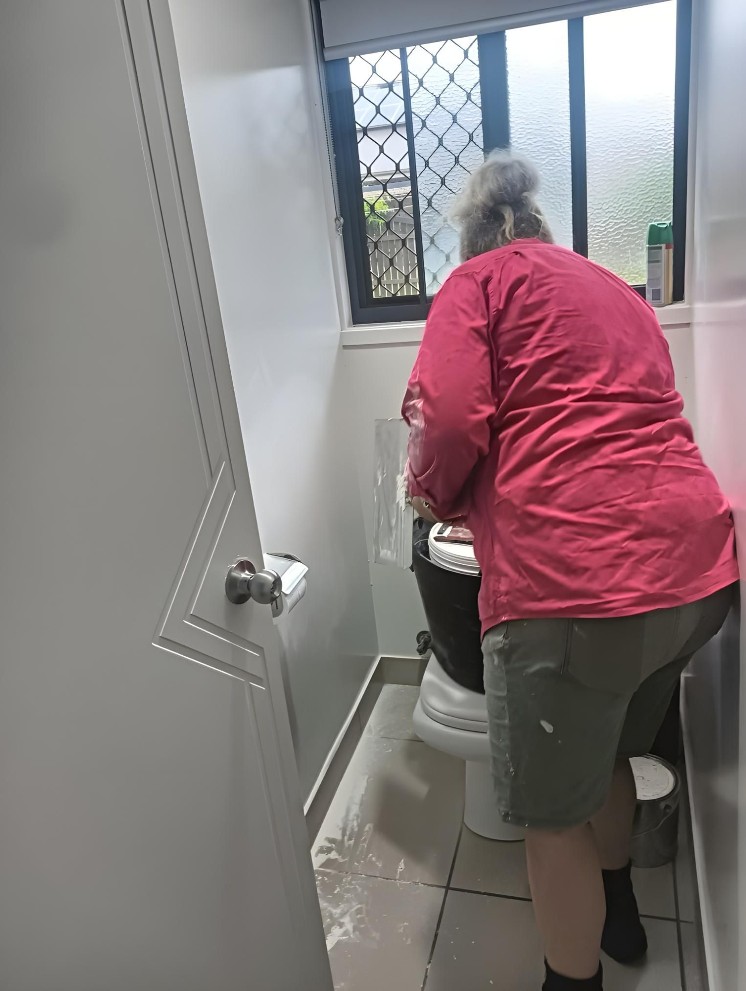 A Woman in a Red Shirt is Standing Next to a Toilet in a Bathroom — Handyman Bundaberg In Burnett Heads, QLD