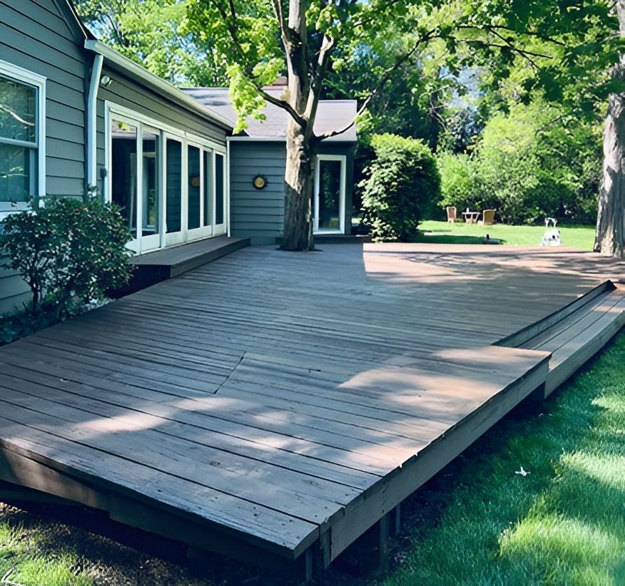 A large, brown wooden deck wraps around a house and tree in a grassy yard.