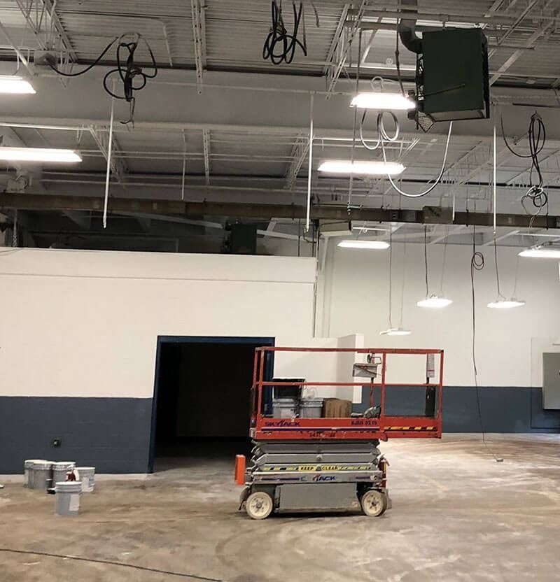 An indoor construction site. A scissor lift is near a wall painted white and gray. Exposed ceiling utilities and wires.