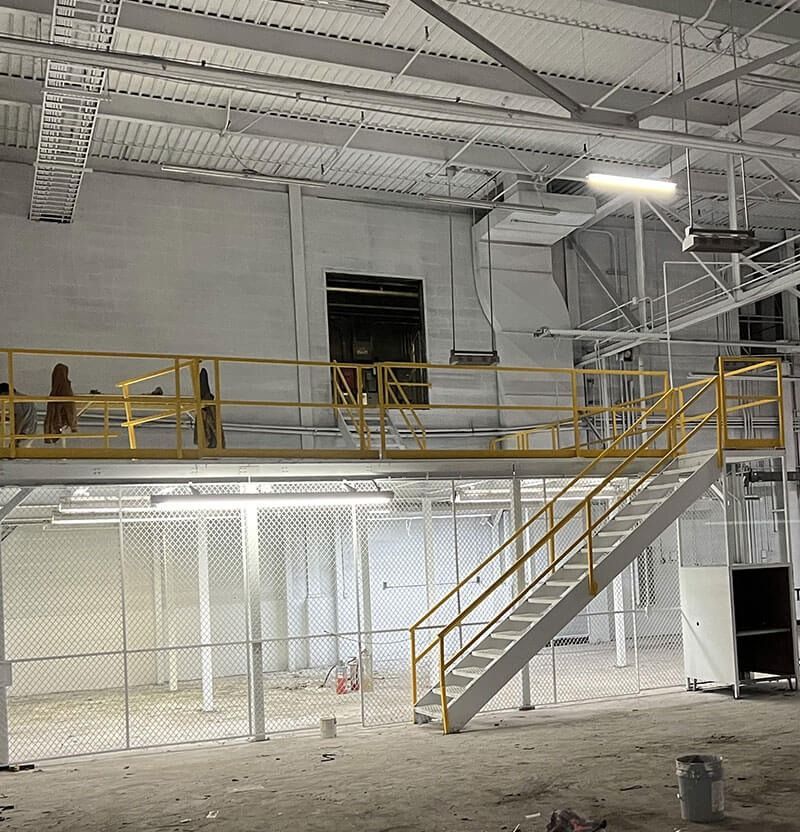 Interior industrial space with a yellow-railed mezzanine, stairs, and wire mesh fencing, painted white.