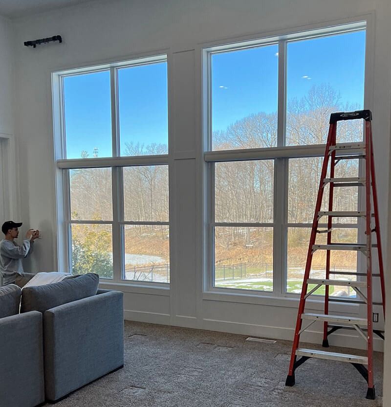 Person installing curtains on a tall window, using a ladder. Interior view with a blue sky outside.