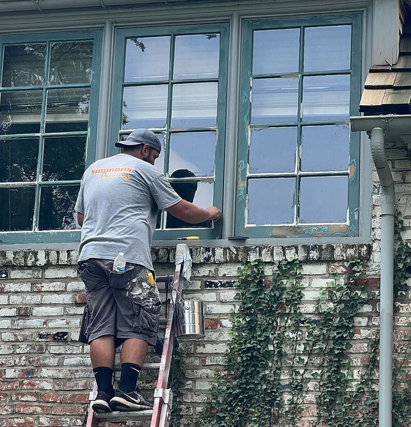 Man on ladder painting window frame outside a brick house; windows are blue-green.